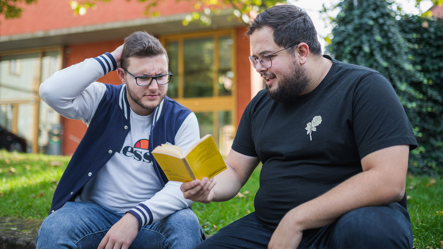 Anton (links) und Dan sitzen im Innenhof des Uni-Geländes und grübeln über einem Buch