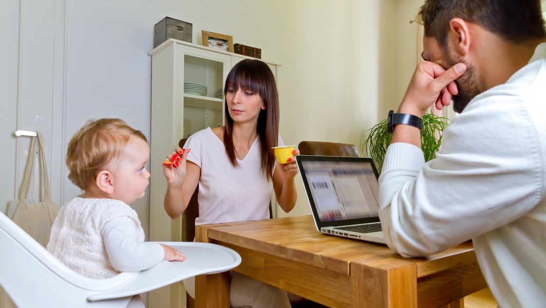 Familie mit Mutter, Vater und Kleinkind sitzen am Tisch. Die Mutter füttert das Kind, der Vater arbeitet am Laptop.