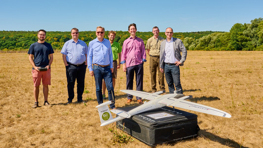 Projektleiter Jonas Troles von der Universität Bamberg, Finanzreferent Bertram Felix, Oberbürgermeister Andreas Starke, Michael Weber vom Sachgebiet Grünanlagen der Bamberger Service Betriebe (BSB), Smart-City-Programmleiter Sascha Götz, städtischer Forstamtsleiter Johannes Hölzel sowie Digitalisierungsreferent Dr. Stefan Goller stehen auf einem Feld vor einer Drohne.