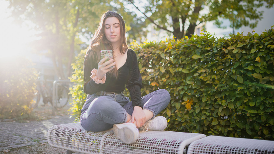 Anna sitzt auf einer Bank und bedient ihr Mobiltelefon