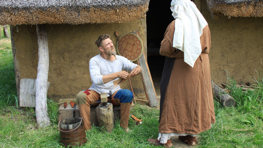 Ein mittelalterliches Paar vor ihrem Haus: er erledigt Handwerk, sie ist in Haushaltsschürze gekleidet.