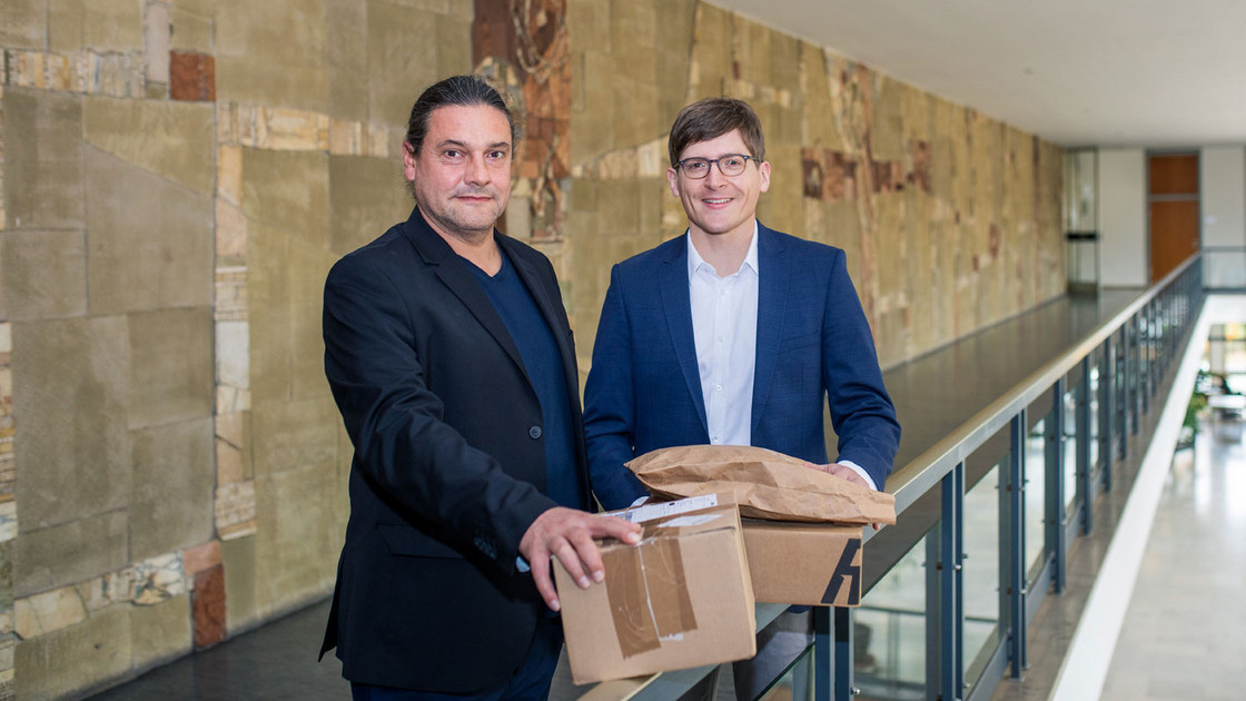 Eric Sucky und Björn Asdecker stehen nebeneinander in der Universität mit Paketen in der Hand