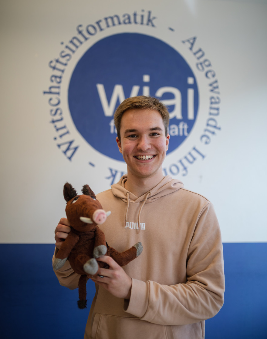 Tobias hält grinsend das Fachschaftsmaskottchen