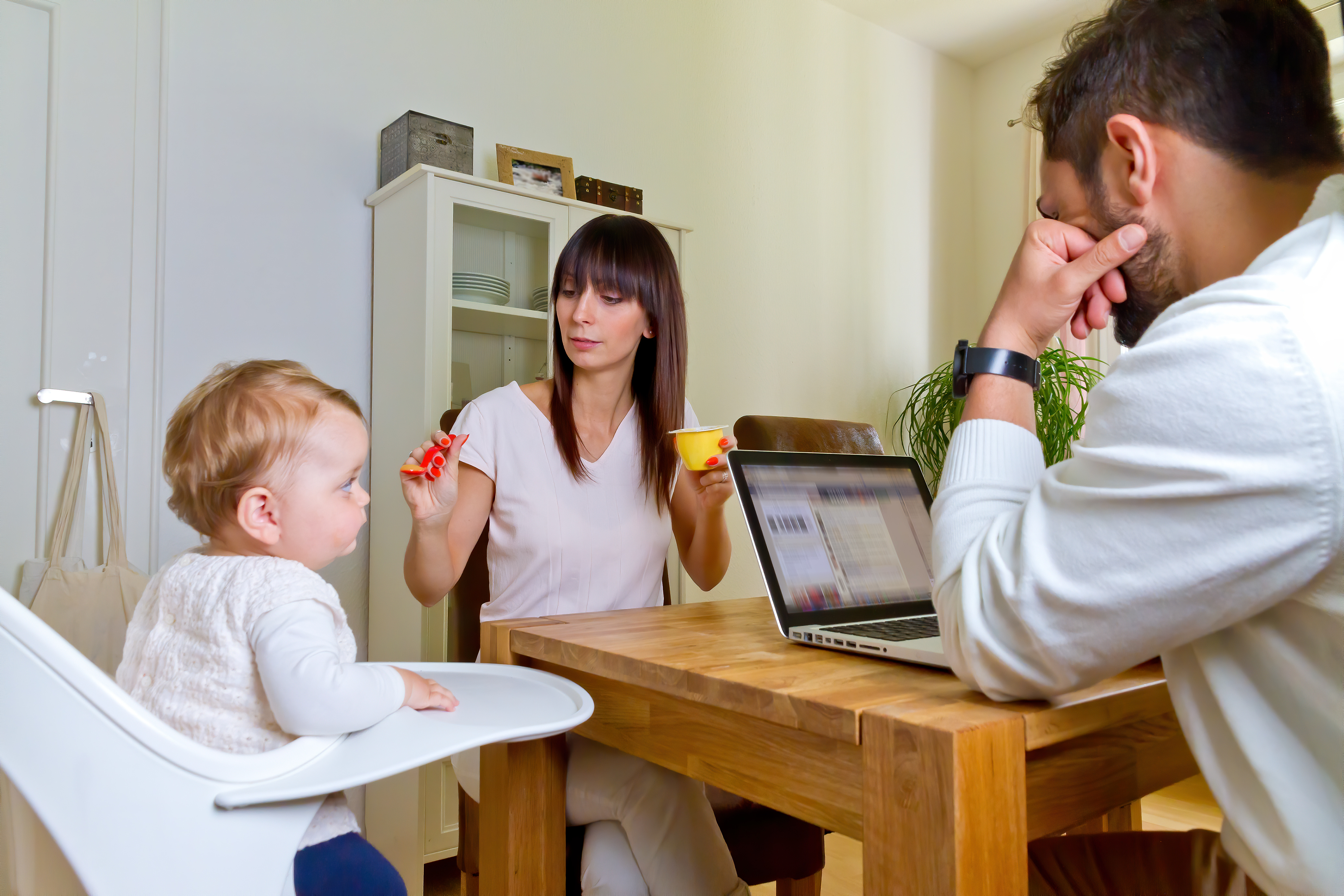 Familie mit Mutter, Vater und Kleinkind sitzen am Tisch. Die Mutter füttert das Kind, der Vater arbeitet am Laptop.